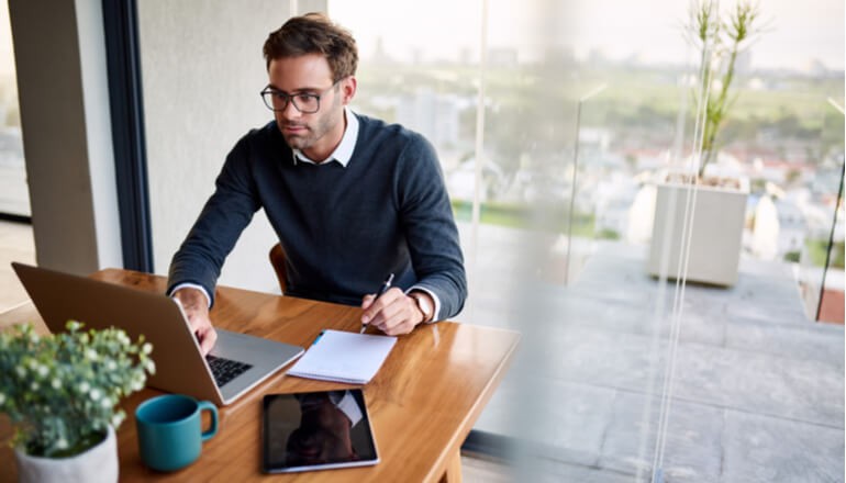 Business professional taking notes while reading report off of laptop